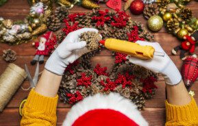 Imagem de mão femininas com luvas brancas fazendo enfeites de natal com uma pistola de cola quente.