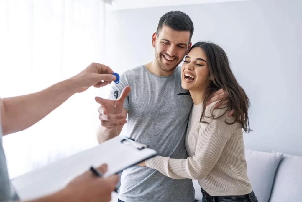 Imagem de um casal sorridente em um apartamento recebendo com entusiasmo um conjunto de chaves de uma pessoa segurando uma prancheta