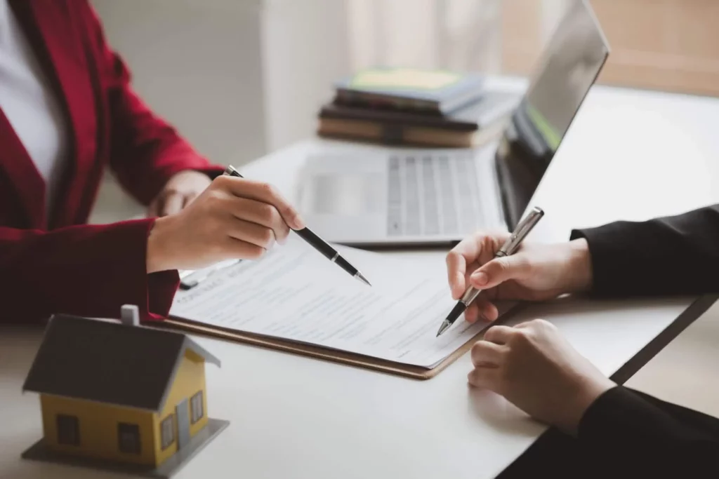 Imagem de duas pessoas sentadas em uma mesa com um laptop e documentos, uma apontando para uma prancheta enquanto a outra assina um formulário