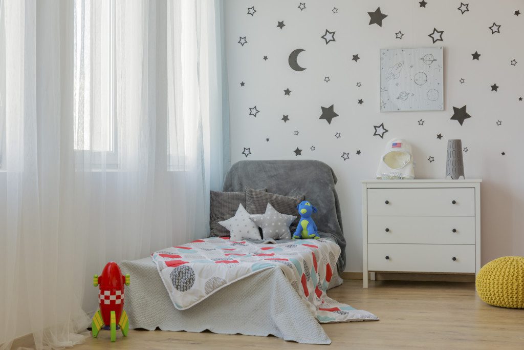  Foto de um quarto com detalhes do espaço sideral. Há uma parede branca com adesivos pretos de estrelas e luas. Tem também uma cama com almofadas de estrelas. Há ainda um foguete de brinquedo e um capacete de astronauta em cima da cômoda.