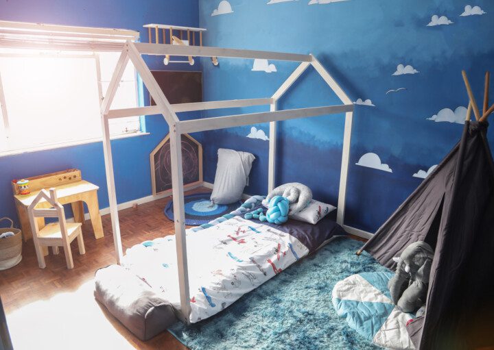 Imagem de um quarto com paredes pintadas de azul. Nelas há nuvens imitando o céu. Nele há também uma cama, uma cabana, almofadas de bichos e uma escrivaninha pequena com cadeira para criança.