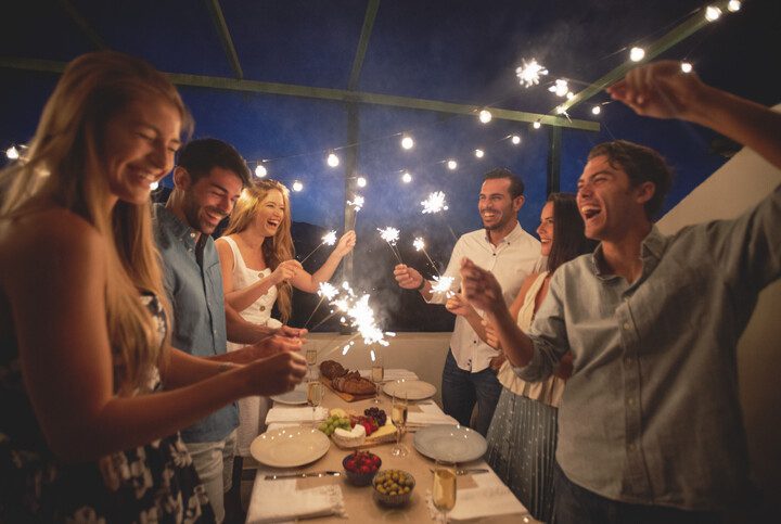 Imagem de um grupo de amigos segurando faíscas, em uma mesa posta de ano novo e festejando.