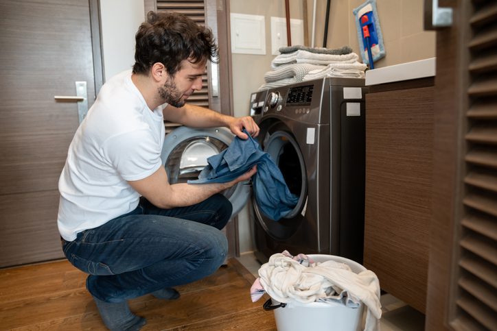 Foto que ilustra matéria sobre consumo consciente de água mostra uma máquina de lavar cheia de roupas e uma mulher, da cintura pra baixo, pegando mais peças em um cesto para colocar na máquina