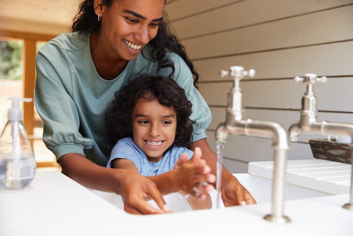 Foto que ilustra matéria sobre consumo consciente de água mostra uma torneira aberta onde aparece em detalhe duas mãos de uma pessoa que se lava.