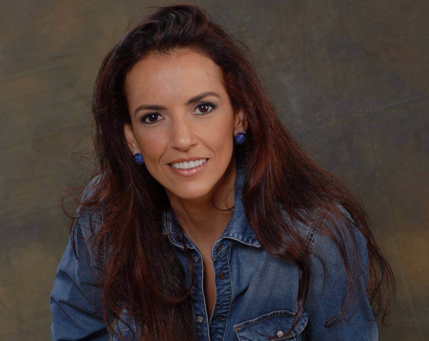 Uma mulher com longos cabelos castanhos, vestindo uma camisa jeans e brincos azuis, sorri para a câmera contra um fundo marrom simples, pronta para compartilhar dicas sobre como decorar o quarto gastando pouco.