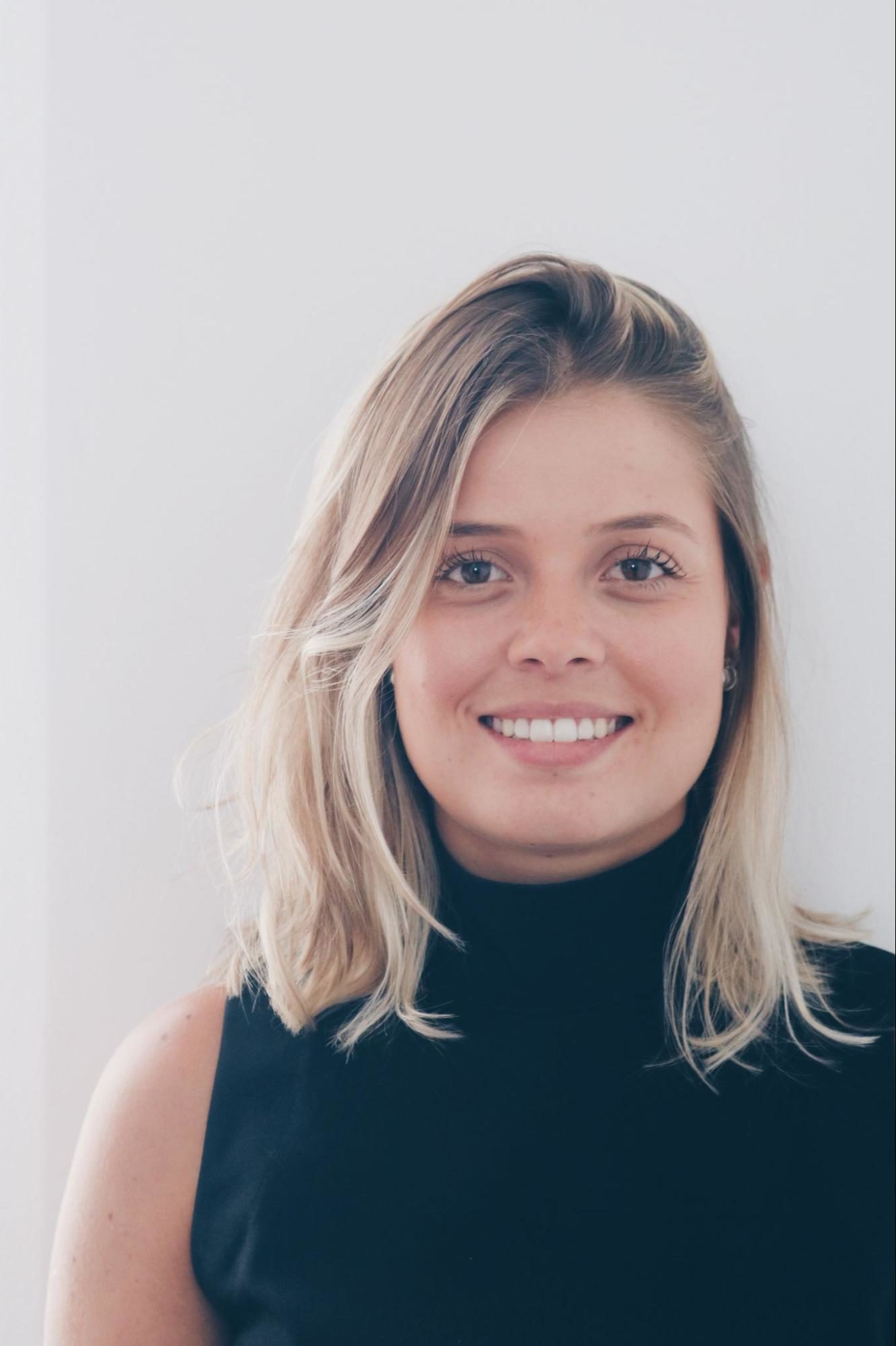 Uma jovem com cabelos loiros na altura dos ombros sorri para a câmera. Vestida com uma blusa preta de gola alta sem mangas, ela posa contra um fundo branco simples, evocando um sutil charme vintage da Quarto.