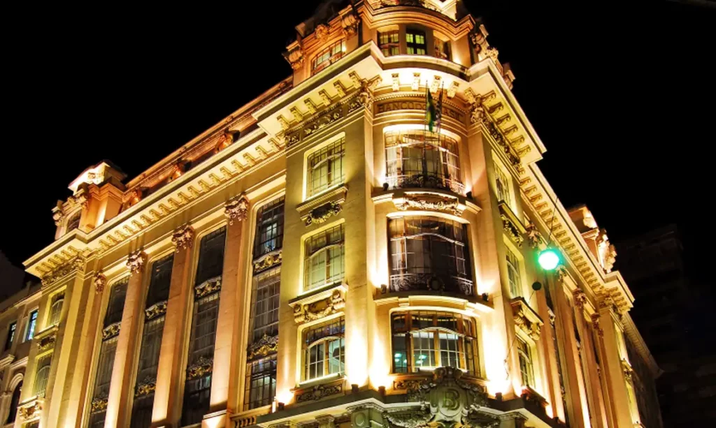 Imagem do edifício histórico onde fica o Centro Cultural Banco do Brasil, CCBB São Paulo. O prédio está iluminado e tem detalhes arquitetônicos ornamentados e colunas altas, fotografado à noite de um ângulo baixo. A iluminação quente destaca desenhos intrincados contra o céu escuro