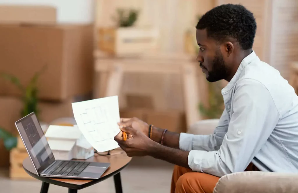 Imagem de um homem sentado em uma pequena mesa, olhando para papéis do ITCMD e segurando um lápis, com um laptop aberto à sua frente. Caixas de papelão e plantas ao fundo sugerem um ambiente doméstico ou de escritório.