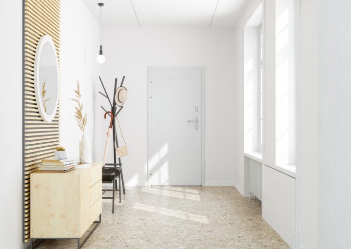 Foto de um corredor localizado na entrada do lar. Nele há um cabideiro com itens pendurados (chapéu, bolsa, sombrinha), um aparador, um vaso com planta e um espelho redondo na parede. O local é bem iluminado por duas janelas grandes.