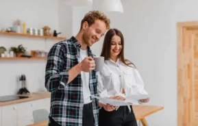 Imagem de um casal formado por um homem e uma mulher em pé na cozinha de um imóvel analisando alguns documentos enquanto o homem toma uma xícara de café para ilustrar matéria sobre imóvel sem entrada
