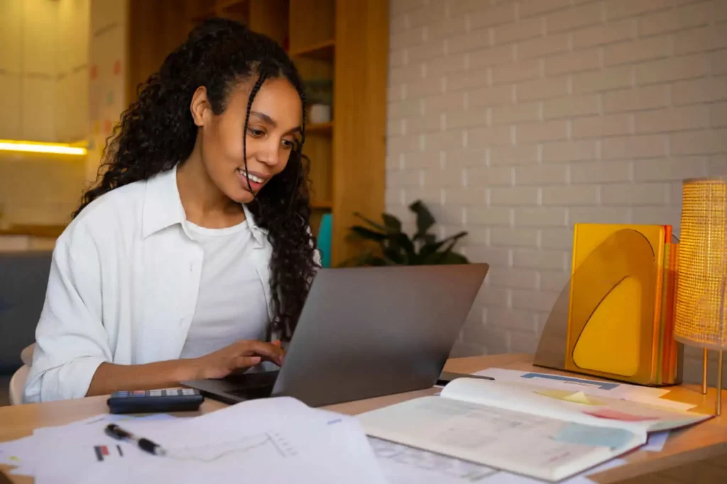 Imagem de uma mulher sentada em uma mesa, sorrindo e trabalhando em um laptop. Há papéis, um notebook, uma calculadora e uma lâmpada sobre a mesa. Ela usa um fone de ouvido, talvez auxiliando em questões do ITCMD, em uma sala moderna com paredes de tijolos brancos.