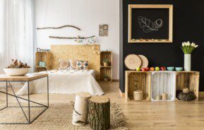 Quarto aconchegante com design rústico inspirado na natureza, com cama de madeira, mesas de cabeceira diferentes, prateleiras de caixotes, troncos decorativos, frutas, tulipas, um desenho de giz de bolota em uma parede preta e móveis em tons neutros.