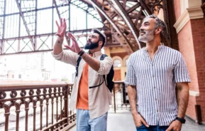Imagem de dois homens em uma estação de metrô em SP. Um deles gesticula animadamente e o outro sorri enquanto a luz do sol passa pelo teto de ferro e vidro acima deles