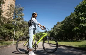 Imagem de uma mulher parada em cima de uma bicicleta com as mãos no guidão olhando para o horizonte em uma ciclovia rodeada por árvores para ilustrar matéria sobre os parques em SP