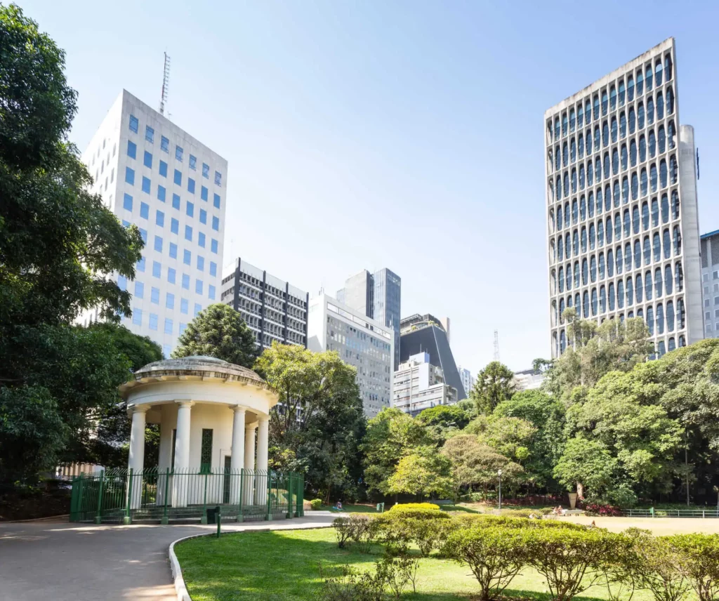 Imagem de parte do Parque Trianon, em São Paulo, mostra gramado, vegetação e infraestrutura do espaço, além de prédios ao fundo, em um dia de céu azul