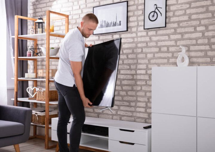 Foto que ilustra matéria sobre quanto custa mobiliar um apartamento mostra um homem segurando uma grande TV de LED para posicioná-la sobre um rack branco em uma sala de estar.