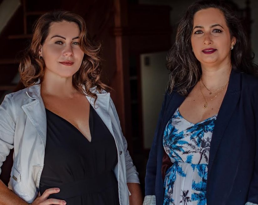 Duas mulheres, lado a lado, com cabelos ondulados, esbanjam confiança em um ambiente interno. Uma usa um blazer branco sobre um vestido preto, enquanto a outra veste um blazer escuro e um vestido estampado azul e branco, ambos emoldurados por paredes setorizadas com pintura suavemente iluminada.