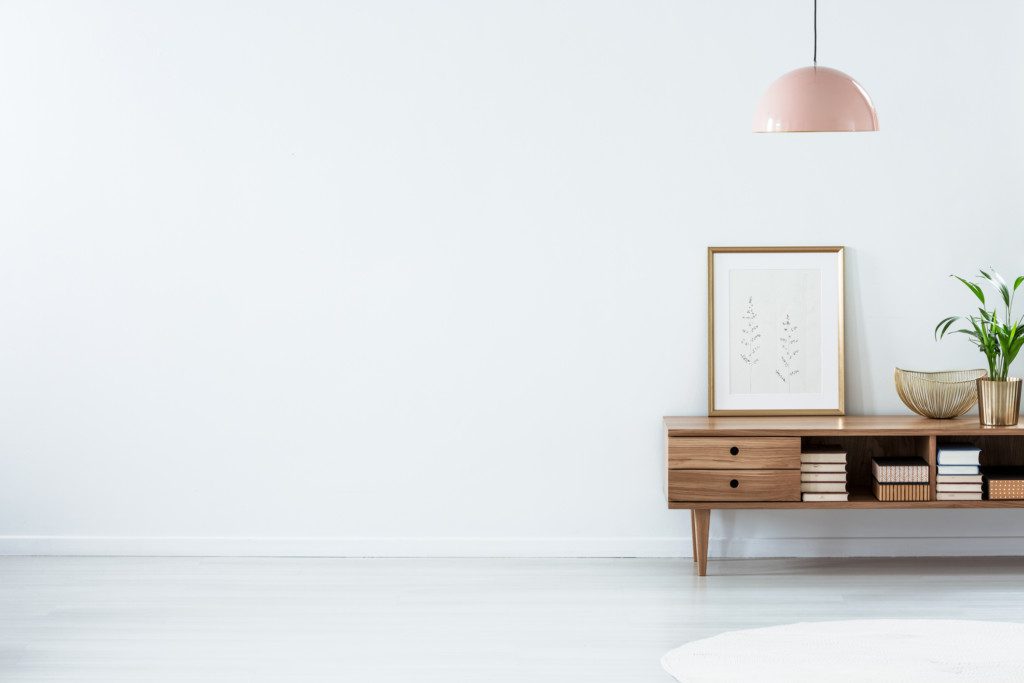 Sala de estar minimalista com um aparador de madeira exibindo decoração com livros, uma obra de arte emoldurada, uma planta e tigelas decorativas. Um pendente rosa está pendurado acima, complementando as paredes e o piso brancos para um visual arejado.