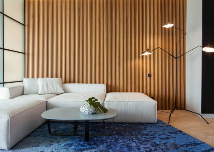 Foto que ilustra matéria sobre decoração com painel ripado mostra uma sala com um sofá branco em cima de um tapete azul. À frente dele há uma mesinha de centro. E atrás, há uma grande parede com um painel ripado de madeira.