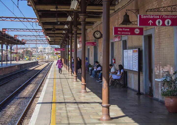 Foto que ilustra matéria sobre a Estação Jundiaí mostra a plataforma da estação de trem em um dia de céu azul, com pessoas sentadas aguardando a chegada do veículo à direita da imagem e os trilhos vazios à esquerda.