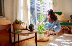 Imagem de uma mulher cuidando de uma planta ao lado de uma mesa com vela e um buda decorativo.