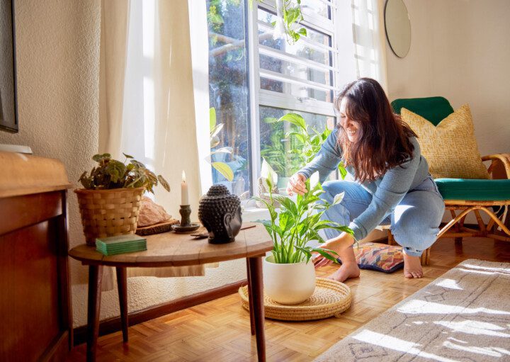 Imagem de uma mulher cuidando de uma planta ao lado de uma mesa com vela e um buda decorativo.