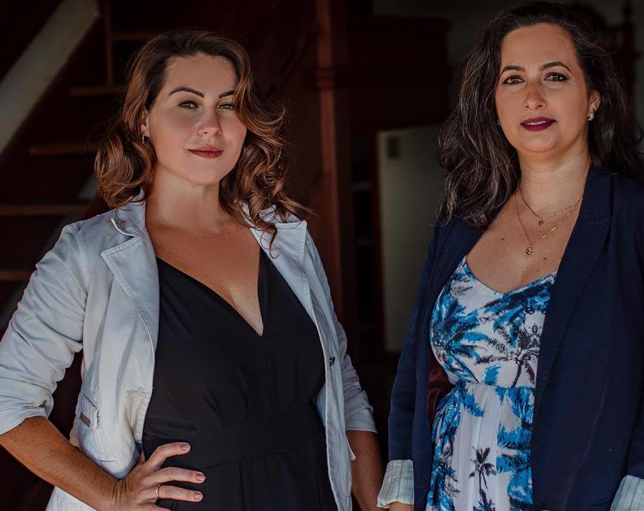 Duas mulheres estão confiantes em um ambiente interno, perto de uma escada, em frente a uma parede geométrica. Uma usa um blazer branco sobre um vestido preto; a outra usa um blazer escuro sobre um vestido estampado azul e branco. Ambas olham diretamente para a câmera.