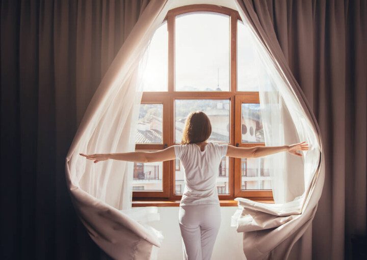 Foto que ilustra matéria sobre tipos de cortina mostra uma mulher de costas abrindo cortinas e olhando por uma janela em um dia de sol.