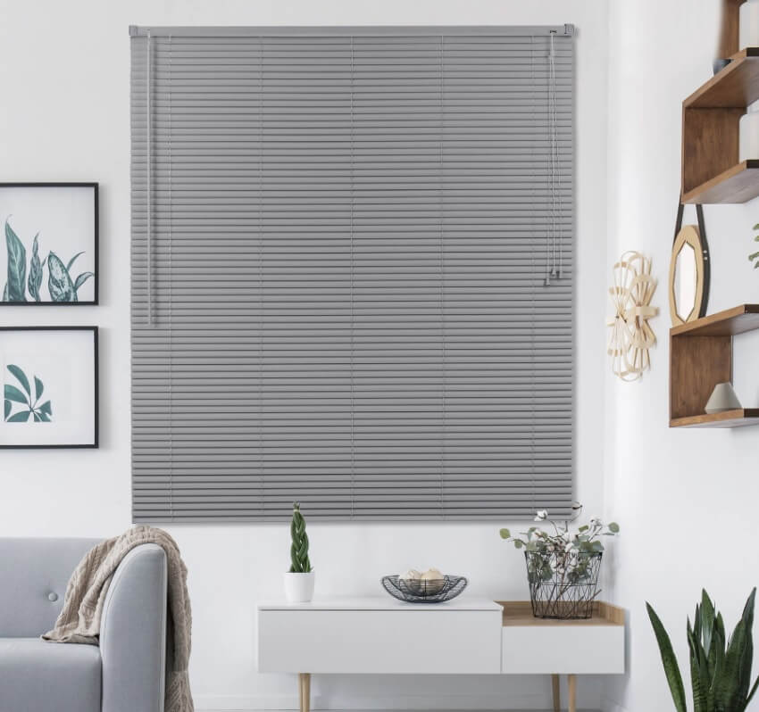 Uma sala de estar moderna apresenta uma grande persiana cinza fechada — um exemplo elegante entre vários tipos de cortina — além de uma mesa de console branca com decoração, arte emoldurada com plantas na parede e prateleiras de madeira com pequenas plantas e acessórios.