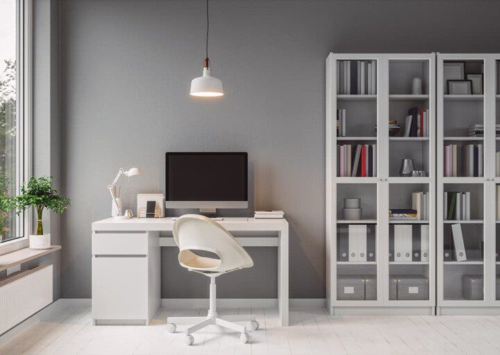 Foto de um escritório com escrivaninha, cadeira, lustre de teto e estante em cor branca. A parede e o computador complementam na cor cinza.