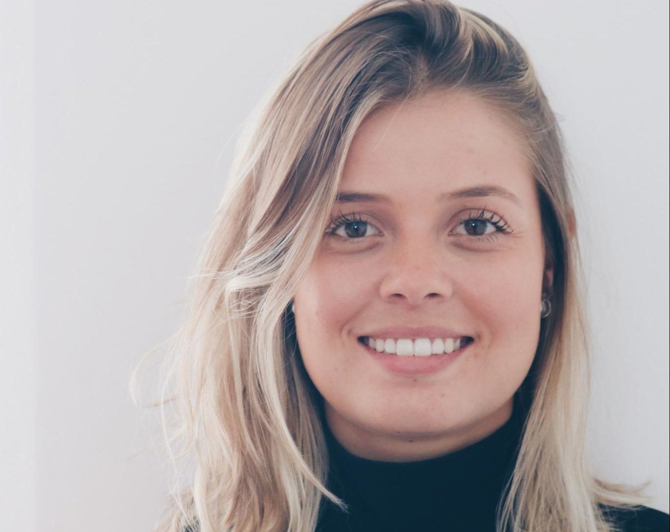 Uma mulher com longos cabelos loiros e uma blusa de gola alta preta sorri para a câmera em frente a um fundo branco simples, evocando o clima limpo e moderno de um escritório minimalista.