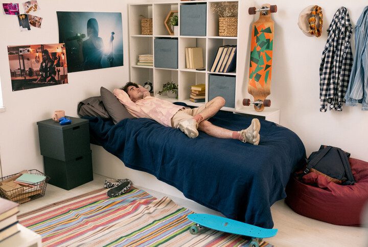 Foto que ilustra matéria sobre quarto geek mostra um homem jovem deitado na cama com posteres nas paredes e um skate pendurado