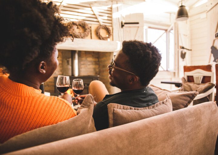 Vista traseira de um jovem casal relaxando no sofá em uma sala de estar rústica, tendo momentos de união e brindando com taças de vinho tinto.