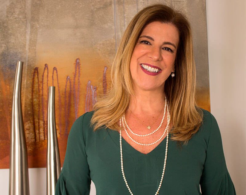 Uma mulher sorridente com cabelos loiros lisos na altura dos ombros usa uma blusa verde e colares de pérolas em camadas, em pé em frente a vasos metálicos modernos e uma pintura abstrata colorida inspirada no Tumblr.