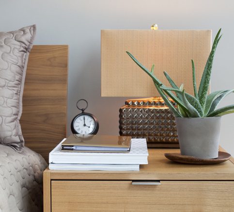 Foto que ilustra matéria sobre decoração para mesa de cabeceira mostra em detalhes uma mesa de cabeceira com um vaso de planta, um abajur e alguns livros empilhados com um relógio despertador em cima.