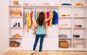 Uma jovem com uma roupa casual está de costas para a câmera, de frente para um grande guarda-roupa branco e tentando escolher roupas para vestir.