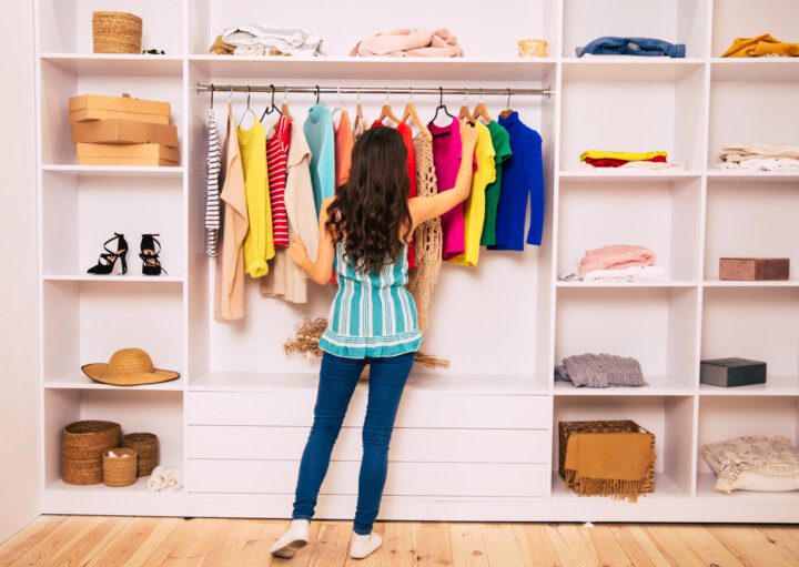 Uma jovem com uma roupa casual está de costas para a câmera, de frente para um grande guarda-roupa branco e tentando escolher roupas para vestir.