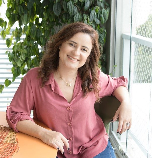 Uma mulher de cabelos castanhos ondulados, vestindo uma blusa rosa e calça jeans azul, está sentada sorrindo em uma mesa perto de uma janela com plantas verdes ao fundo, lembrando o clima aconchegante de um quarto de bebê retrô.