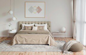 Um quarto romântico moderno e iluminado, com cama bege, cabeceira capitonê, criados-mudos minimalistas, tapete geométrico, pufe, arte abstrata na parede e uma grande janela com cortinas transparentes. A decoração é neutra, com detalhes simples e elegantes.