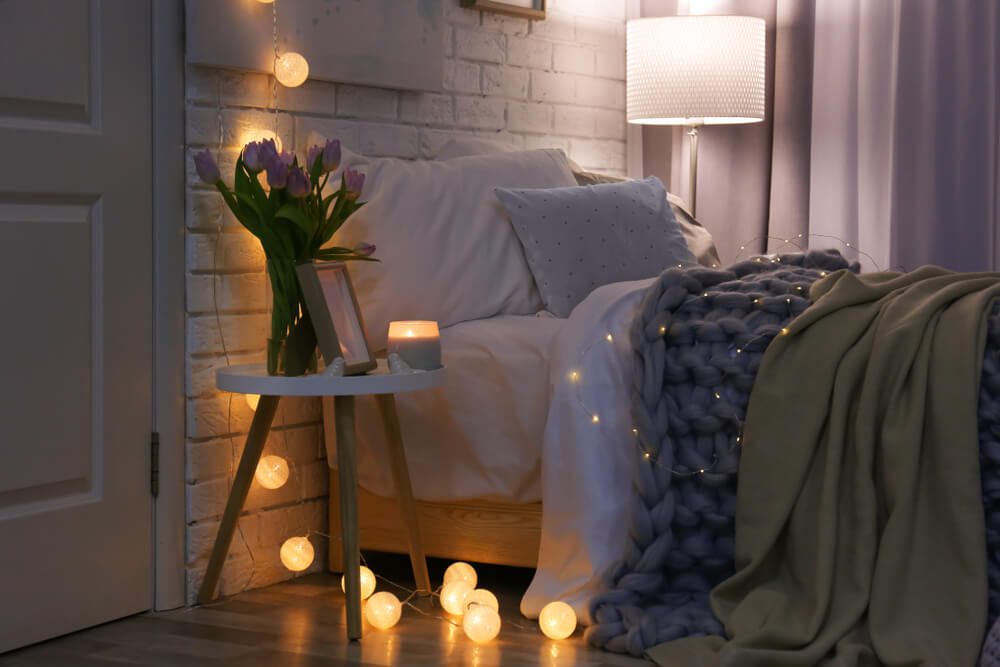 Uma cena Tumblr aconchegante em quarto com uma cama coberta por cobertores grossos, uma mesa de cabeceira com uma vela, um vaso de tulipas roxas e luzes de corda iluminando suavemente o ambiente. Um abajur sobre a mesa emite um brilho aconchegante.