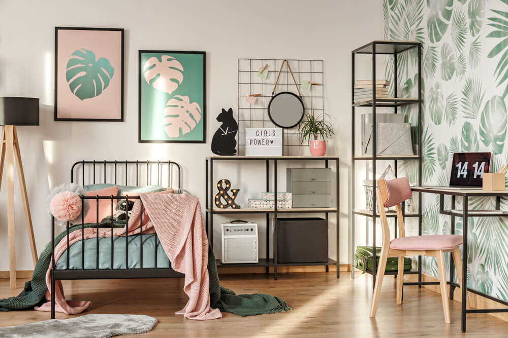 Um elegante quarto Tumblr com uma cama de metal preta, roupa de cama rosa pastel e verde, arte de parede com folhas tropicais, prateleiras com itens de decoração, uma escrivaninha de madeira com uma cadeira rosa e papel de parede com folhas em uma parede.
