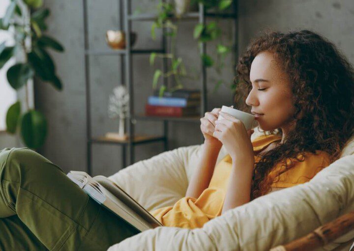 Foto que ilustra matéria sobre sala de descanso mostra uma jovem mulher sentada em uma poltrona com um livro no colo e tomando uma bebida em uma xícara com os olhos fechados