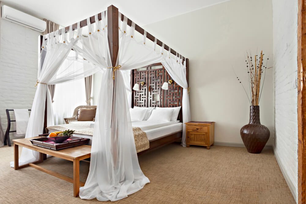 Um quarto moderno tem uma cama de dossel — um dos elegantes tipos de cama — coberta com cortinas brancas, um banco de madeira aos pés com uma bandeja de frutas, um criado-mudo e um grande vaso decorativo com galhos altos no canto.