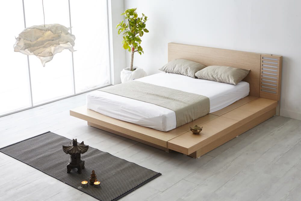 Quarto minimalista com uma moderna cama baixa de madeira, exibindo um dos populares tipos de cama, com roupa de cama bege, uma planta em vaso, uma luminária pendente suave e um tapete de tecido sobre um piso de madeira clara.