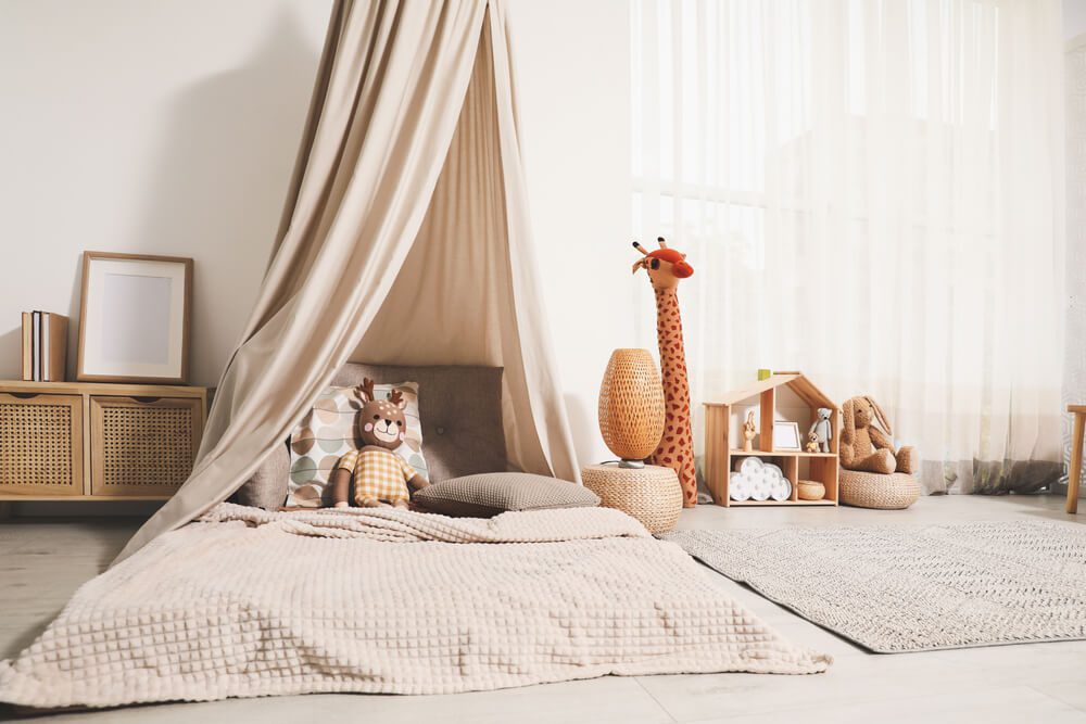 Uma aconchegante sala de jogos infantil com um colchão macio bege no chão sob um dossel de tecido, brinquedos de pelúcia de animais, um armário de vime, uma girafa de brinquedo e um tapete sobre piso de madeira clara exibe um dos encantadores tipos de cama para crianças.