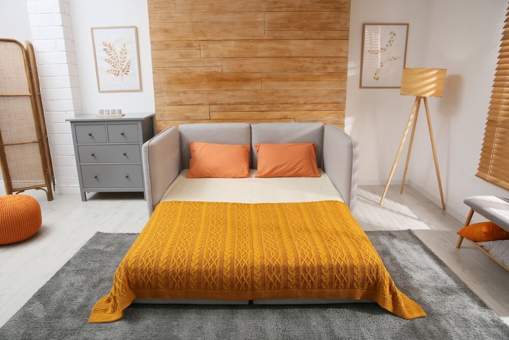 Um quarto aconchegante conta com um sofá-cama cinza, um dos muitos tipos de cama, com almofadas laranja e um cobertor laranja estampado, uma cômoda cinza, quadros, uma luminária de chão e detalhes em madeira, tudo sobre um tapete cinza.
