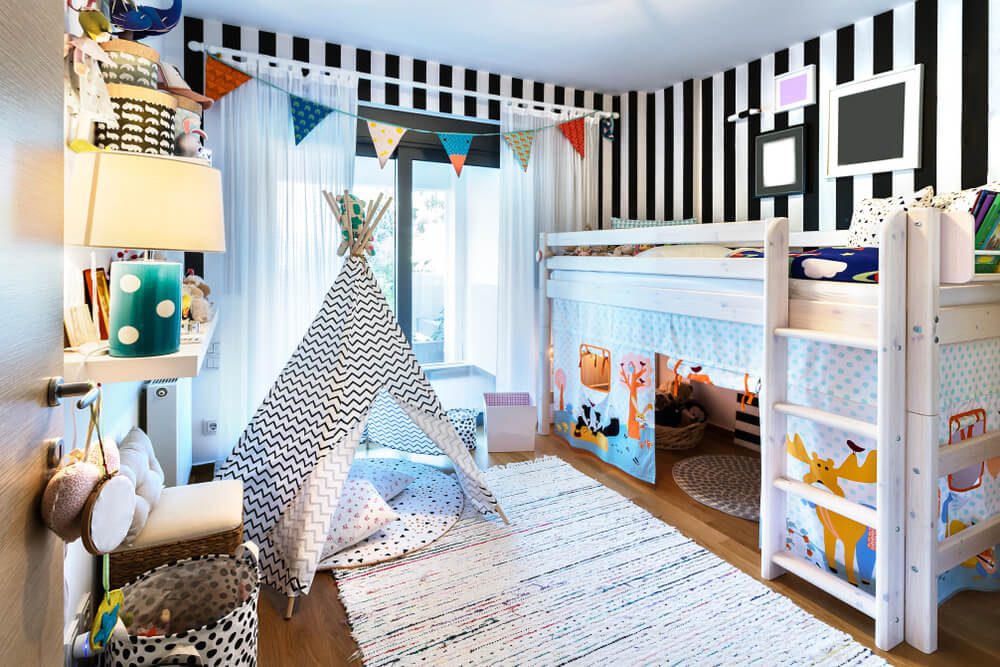 Quarto infantil colorido com beliche, barraca de brinquedo e tapete estampado. O quarto tem paredes listradas em preto e branco, decorações vibrantes, brinquedos, livros e bandeirinhas de barbante perto da janela — exibindo divertidos tipos de cama em um ambiente lúdico.