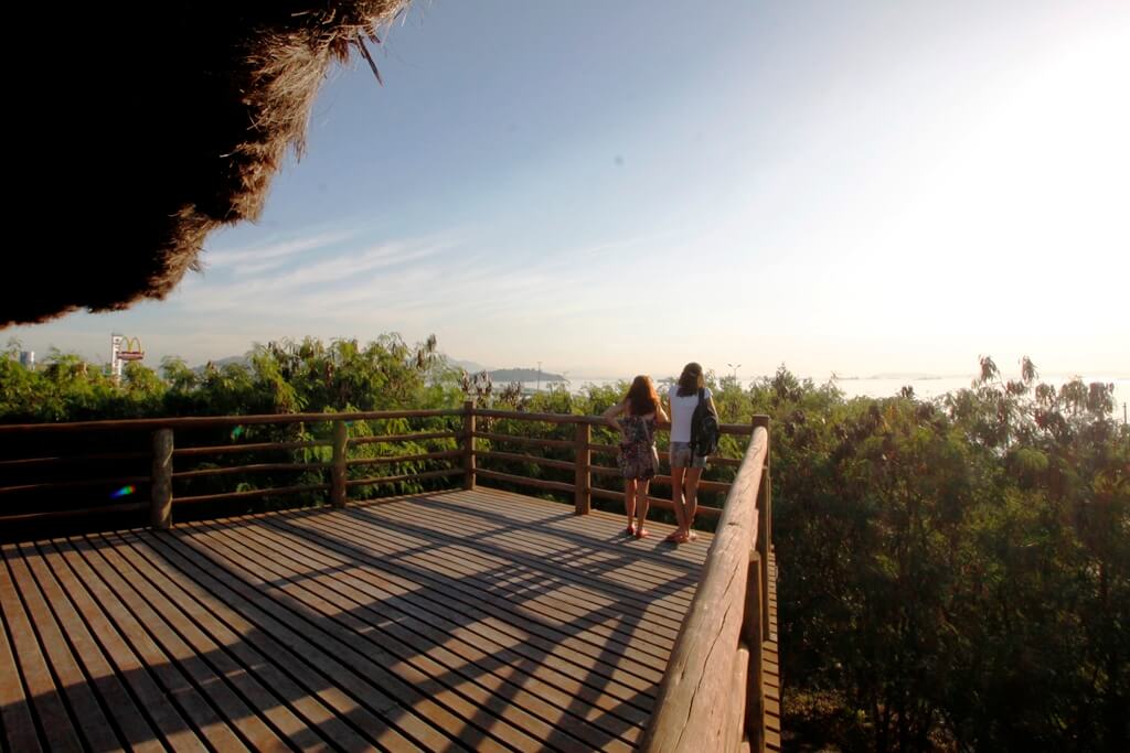 Foto que ilustra matéria sobre os bairros de São Gonçalo mostra duas mulheres de costas em um mirante de madeira olhando para árvores logo abaixo e, ao fundo, a Baía de Guanabara.