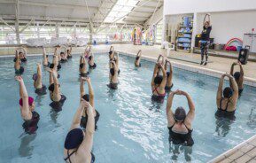 Foto que ilustra matéria sobre o programa Curitiba em Movimento mostra uma piscina com várias mulheres dentro com os braços levantados acima da cabeça fazendo alongamento, enquanto são orientadas por uma instrutora de pé na borda, do lado de fora.