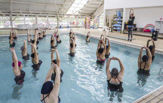 Foto que ilustra matéria sobre o programa Curitiba em Movimento mostra uma piscina com várias mulheres dentro com os braços levantados acima da cabeça fazendo alongamento, enquanto são orientadas por uma instrutora de pé na borda, do lado de fora.